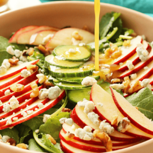 Close up shot of a baby kale and apple salad with blue cheese crumbles and walnuts in a shallow pink bowl with apple cider vinaigrette being poured over it