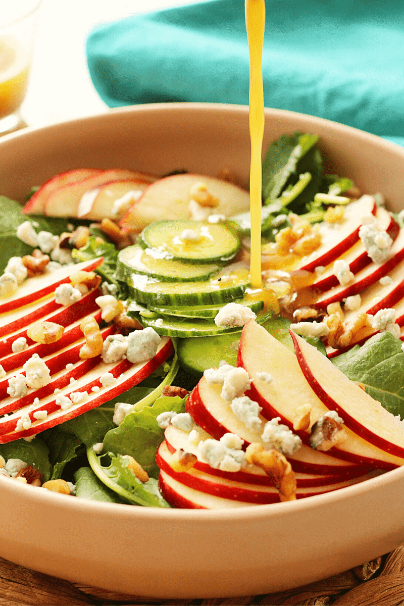 Close up shot of sparkling apple cider vinaigrette being poured over a Baby Kale and Apple Salad in a shallow pink bowl