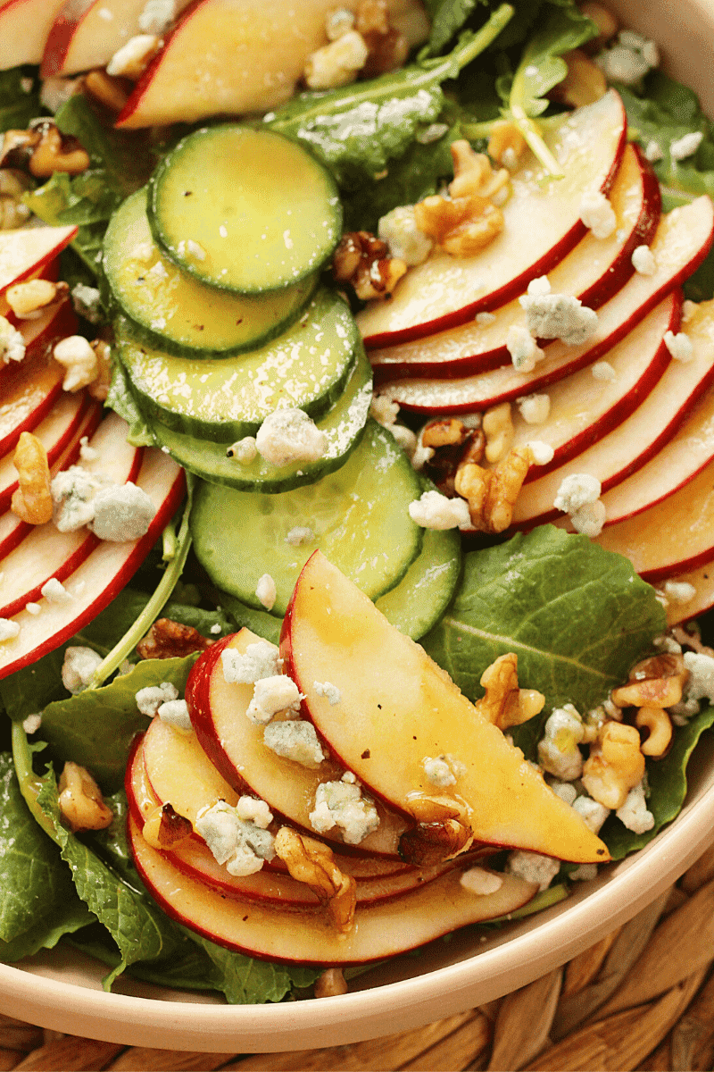 Close up of a Baby Kale and Apple Salad with blue cheese crumbles, walnuts, cucumber rounds, drizzled with apple cider vinaigrette