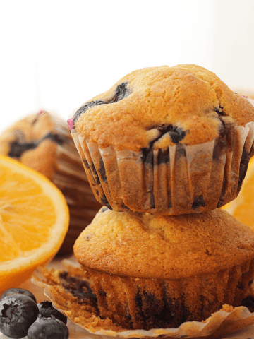 Close up of 2 blueberry muffins stacked on top of the other, blueberries in the foreground and muffin and lemons in the background