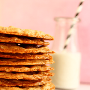 45 degree angle close up of a stack of thin chocolate chip cookies and a glass of milk in the background