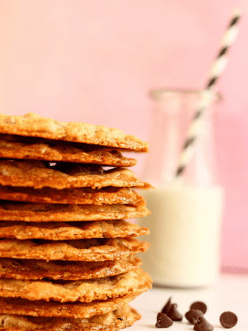 45 degree angle close up of a stack of thin chocolate chip cookies and a glass of milk in the background