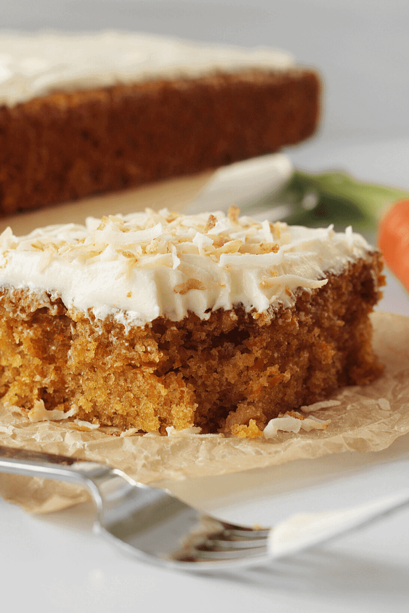 Eye-level close-up of a fluffy carrot cake slice topped with toasted coconut flakes with a fork in foreground. Purpose of image is to show texture.