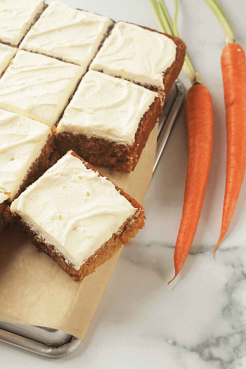 This is an angled overhead shot of a flluffy carrot-sheet cake that's been iced with whipped-cream cheese frosting and cut into squares. The purpose f the image is to suggest to the reader how to slice and serve the cake.