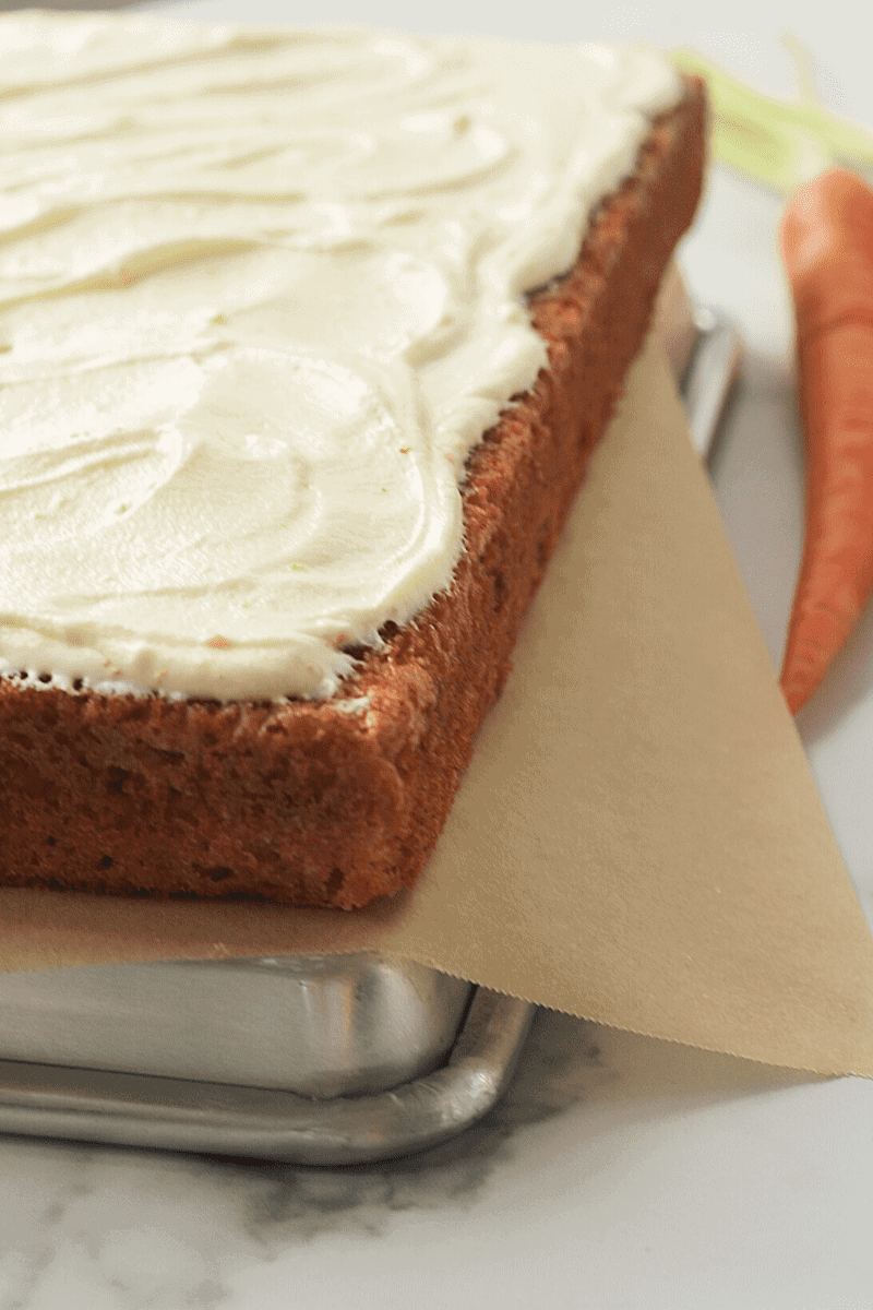 This is a 45 degree close-up of a carrot sheet cake that's topped with cream cheese frosting, displayed on an upside-down sheet pan. This is a close up view meant to entice the reader.