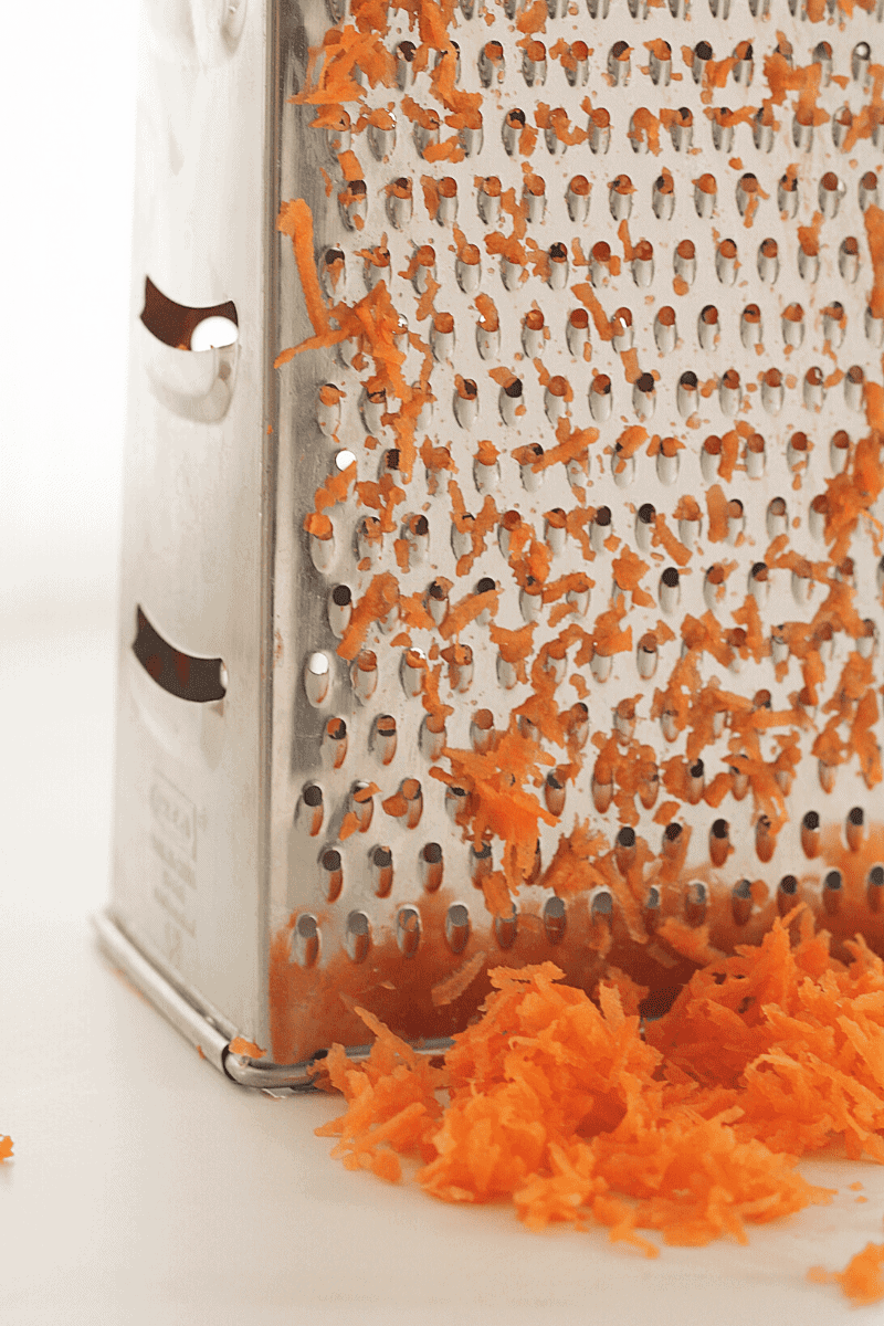 This is an eye-level close-up of carrots on a grater. The purpose of the image is to show the reader what side of the grater they should use (the smaller size) and to get a visual of how fine the carrots should be shredded.