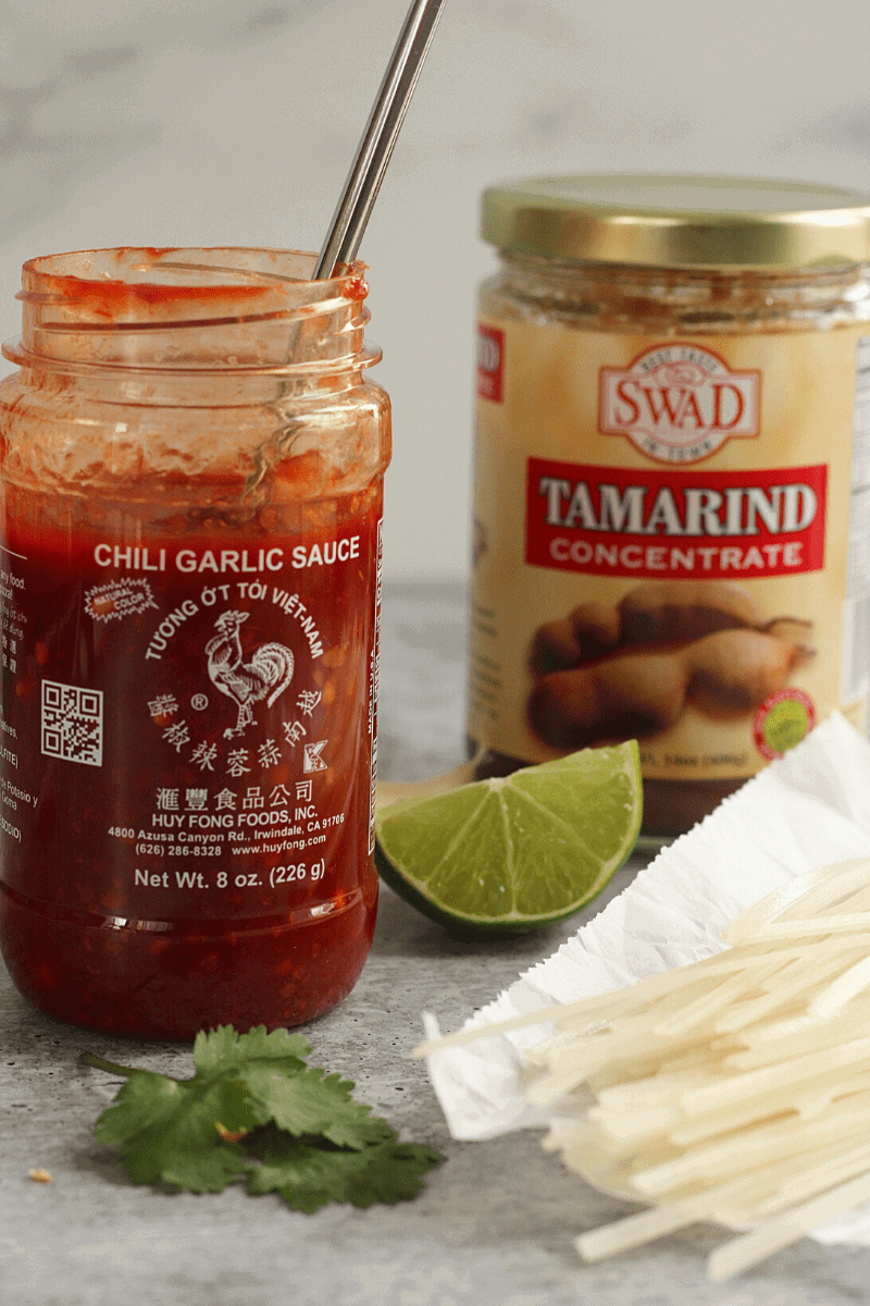 This is a 45 degree close up of jars of chili-garlic sauce, tamarind concentrate, a lime wedge and rice noodles. All key ingredients for this pad thai recipe