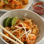 An angled overhead shot of pad thai with shrimp, tofu and green onions in a round bowl with a platter of more pad thai and a small ramekin of chili-garlic sauce in the background. This photo is intended to entice the reader and suggest how to plate and serve the dish.