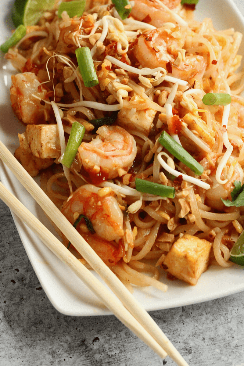 An angled overhead shot of pad thai with shrimp, tofu and green onions on a square plate with chopsticks. This photo is intended to entice the reader and suggest how to plate and serve the dish.