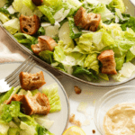 This is a close up of a caesar salad made with romaine lettuce leaves, whole grain croutons and shaved parmesan tossed in homemade caesar dressing. The purpose of the image is to entice the reader and suggest how to plate and serve.