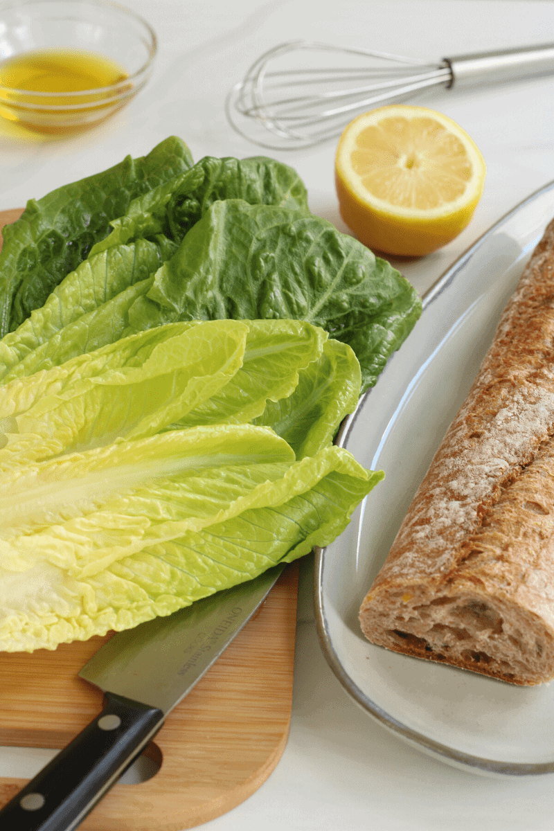 close up of some basic ingredients needed to make a simple Caesar salad: Romaine hearts, lemon, olive oil and a whole grain baguette for the croutons. Not all ingredients for the salad are shown