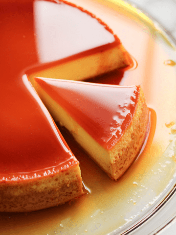 Overhead shot of sliced Cape Verdean pudim (flan) on a clear plate topped with glossy caramel syrup. The purpose of this image is to entice and show the reader how to slice the reader.