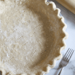 This is a close-up overhead shot of an unbaked, all-butter pie shell that's been docked. There is a wooden rolling pin and a fork off to the side.