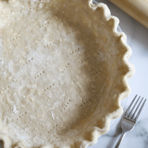 This is a close-up overhead shot of an unbaked, all-butter pie shell that's been docked. There is a wooden rolling pin and a fork off to the side.