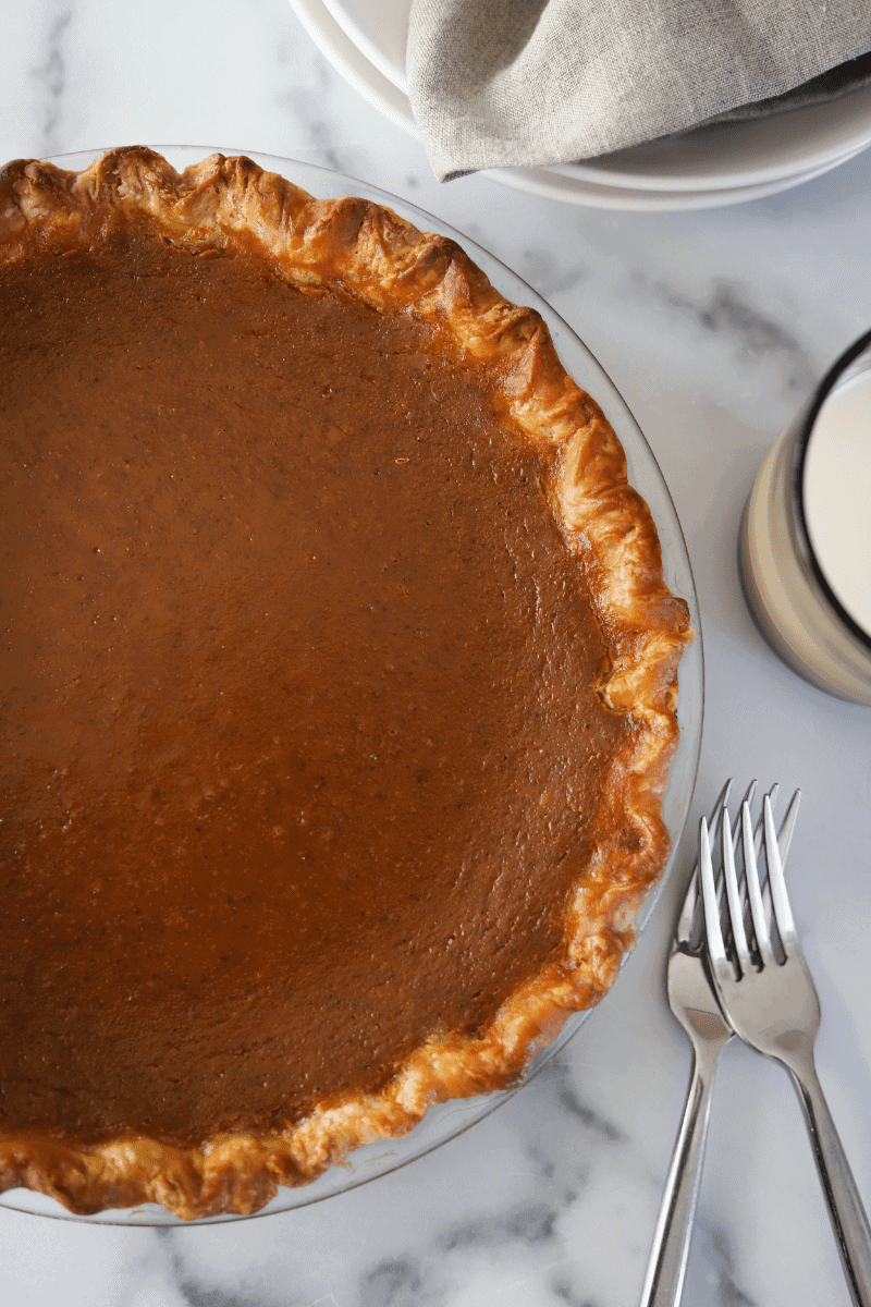 overhead photo of a fully baked pumpkin pie with dishes, a napkin and a glass of milk arranged on the side