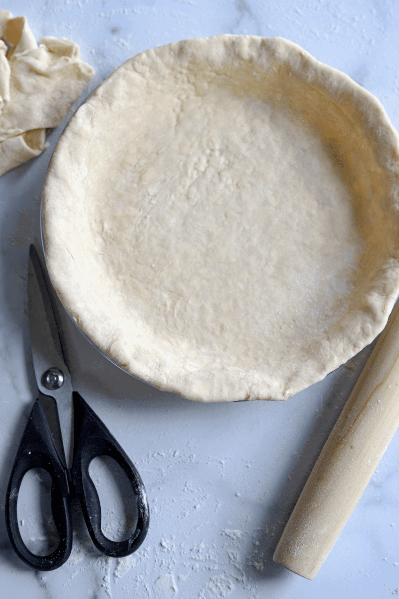 This is an overhead shot of an unbaked all-butter pie crust with scraps of dough and a pair of kitchen scissors off to the side.
