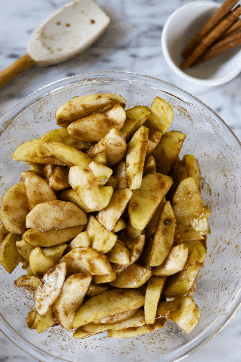 This is a photo of ¼ inch sliced apples tossed in sugar and spices in a bowl. There is a silicone spatula and cinnamon sticks in the background.