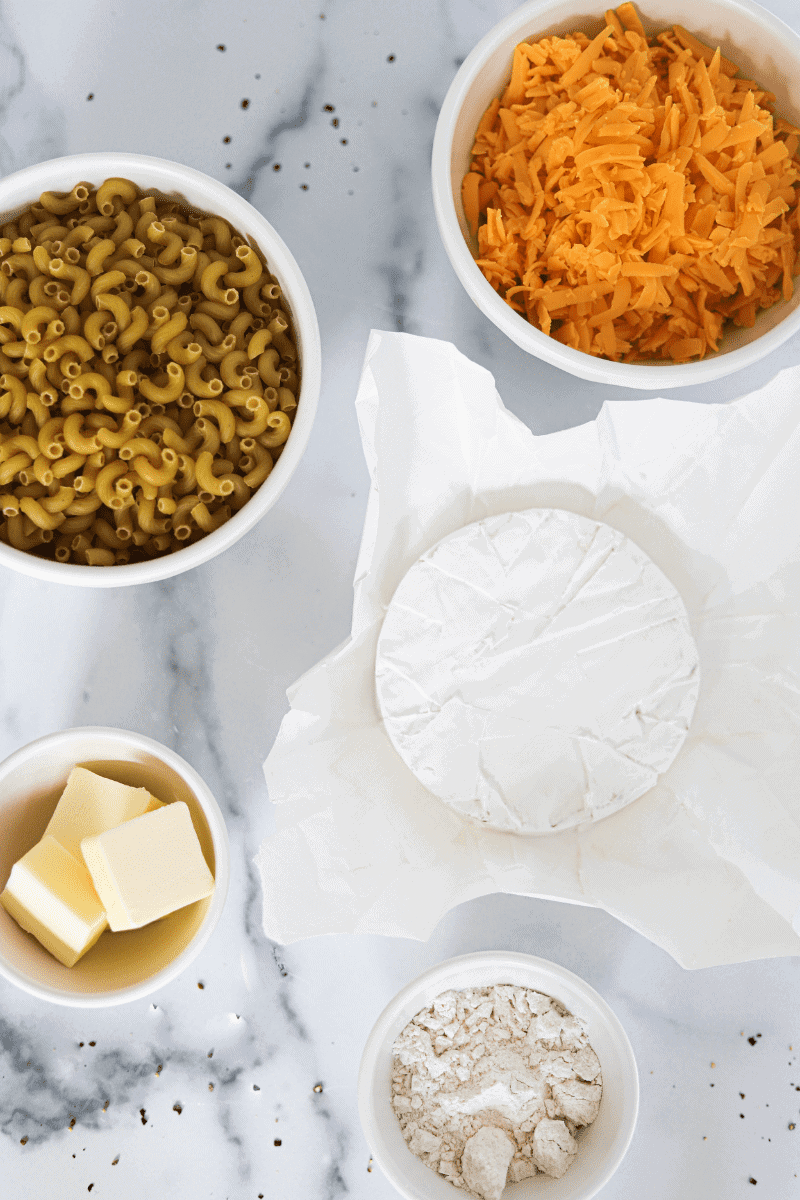 shot of some key ingredients used to make brie and cheddar baked and mac and cheese-shredded cheddar, elbow macaroni, cubed butter, a round of brie cheese and flour