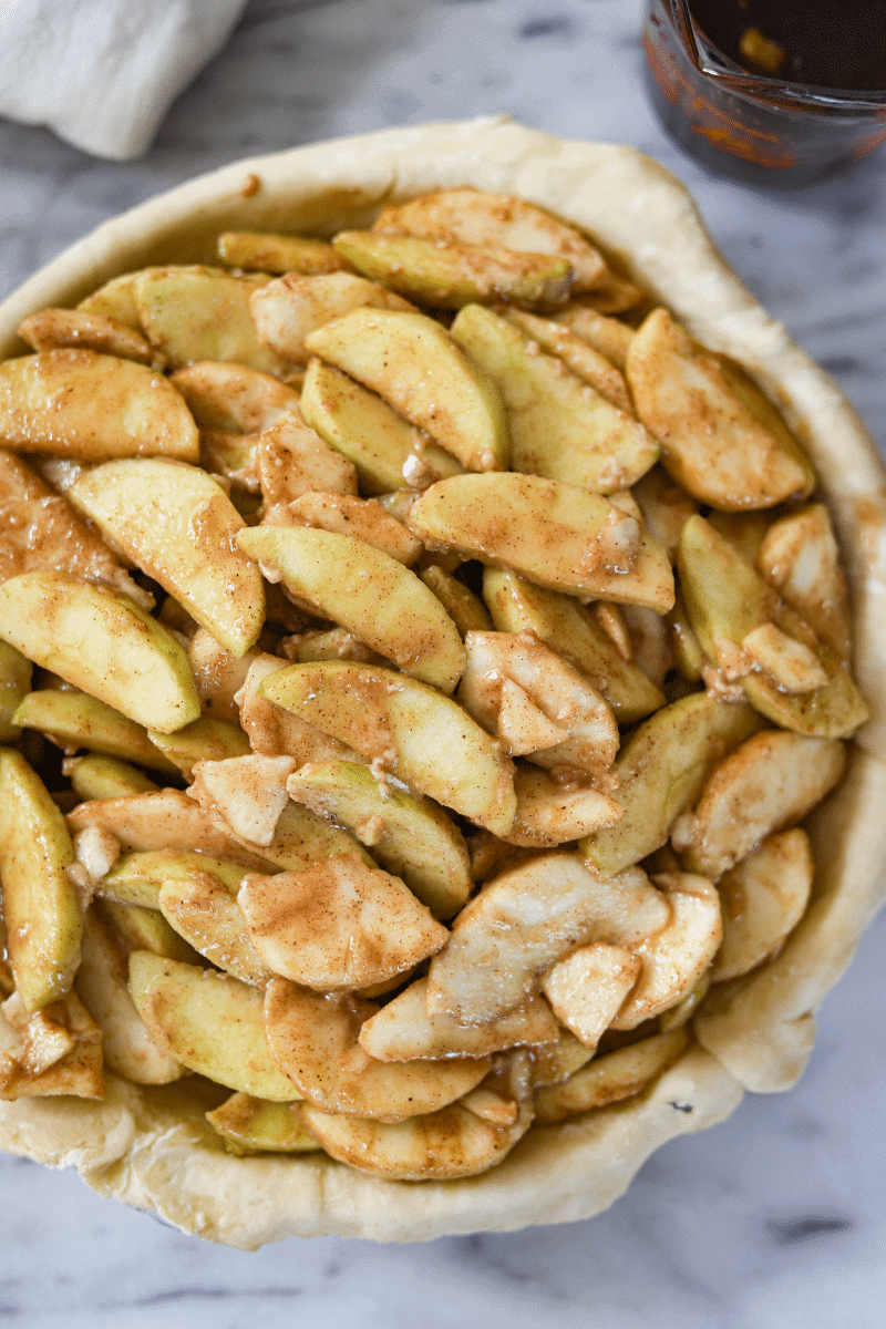 This is a close-up photo of sliced apples coated in sugar, flour and spices, and piled into an unbaked pie shell. The cooked, drained liquid from the apples in the background, and has not been poured over the apples yet.