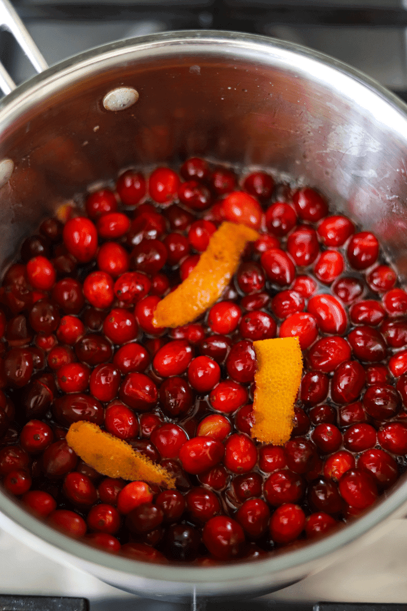 Process shot of fresh cranberries and orange peels in a pan to show the reader how to make spiked homemade cranberry sauce