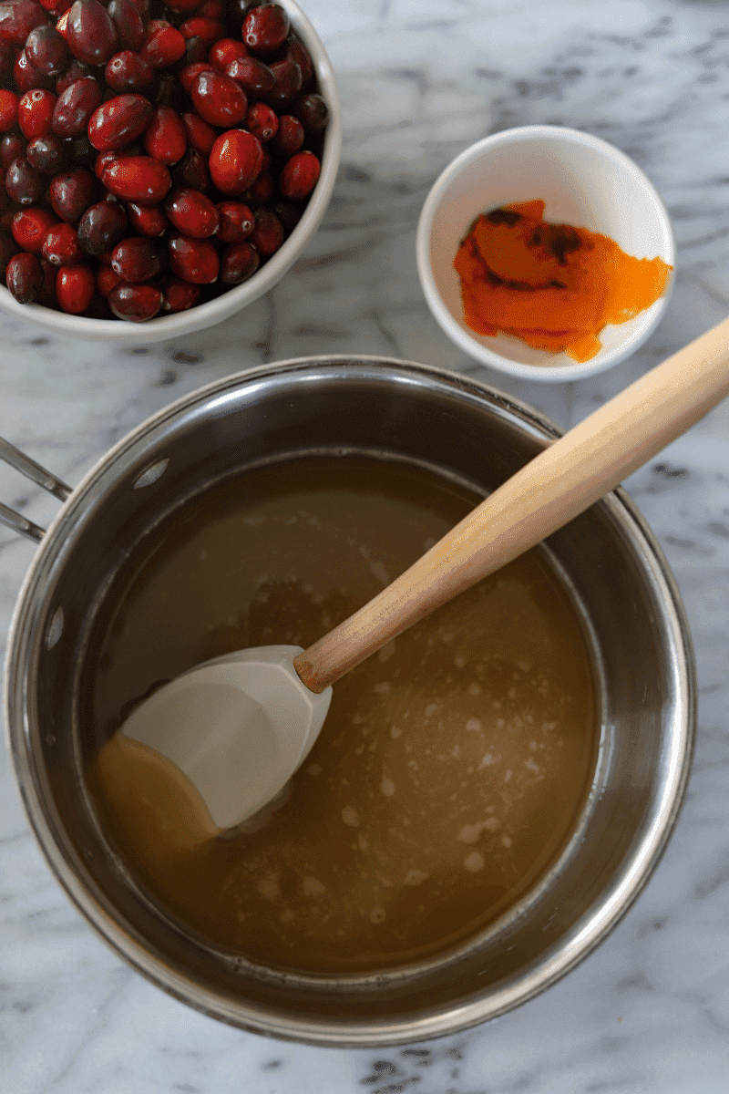 process shot showing brown sugar, triple sec and orange juice in a deep saucepan with fresh cranberries and orange peels in the background. The image is showing the reader how to make spiked homemade cranberry sauce