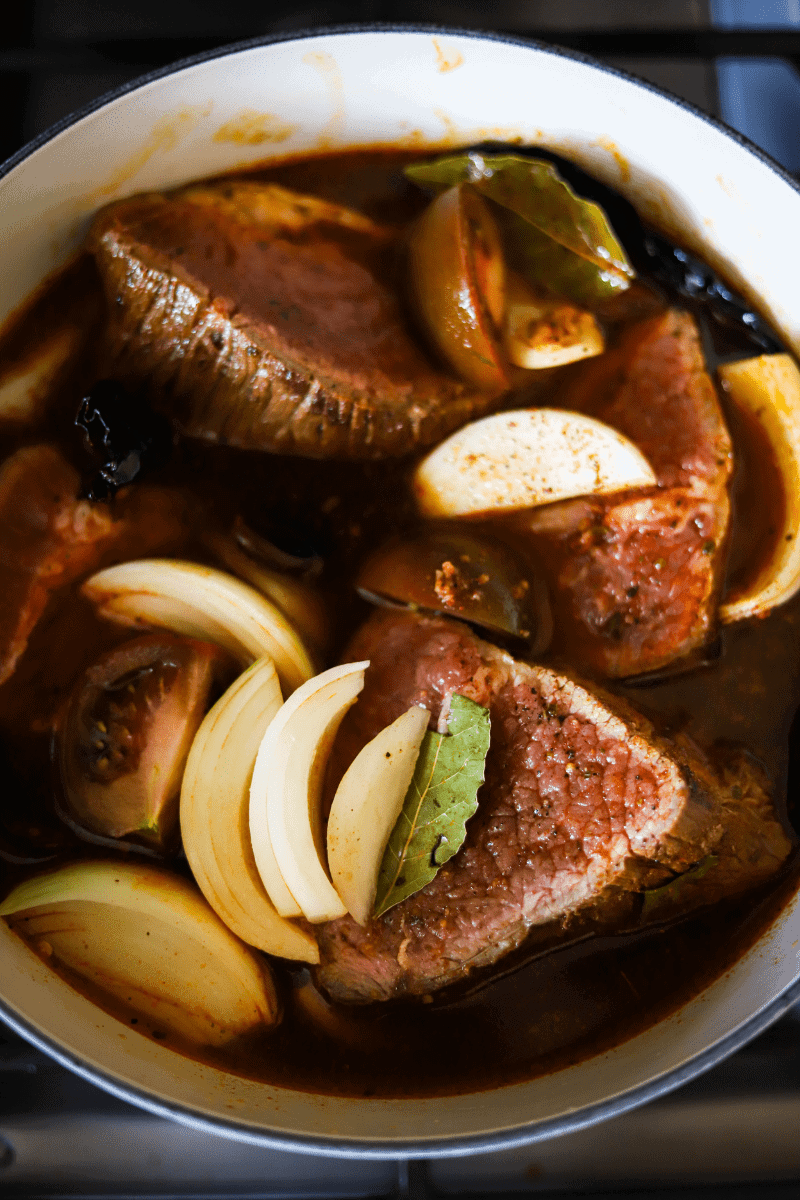 A soup pot filled with guajillo and pasilla peppers, beef roast, quartered onions and tomatoes in preparation for making Mexican birria