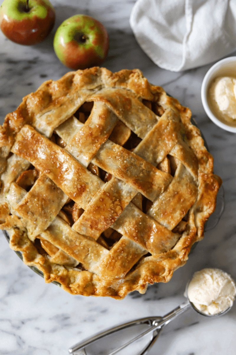 This is a beautiful overhead shot of the fully baked classic apple pie in the center of the photo. A white dish towel is draped off to the side, a metal ice cream scoop, a small bowl of ice cream and a few loose apples are placed strategically to frame the photo. The lattice crust is deep golden brown in color.