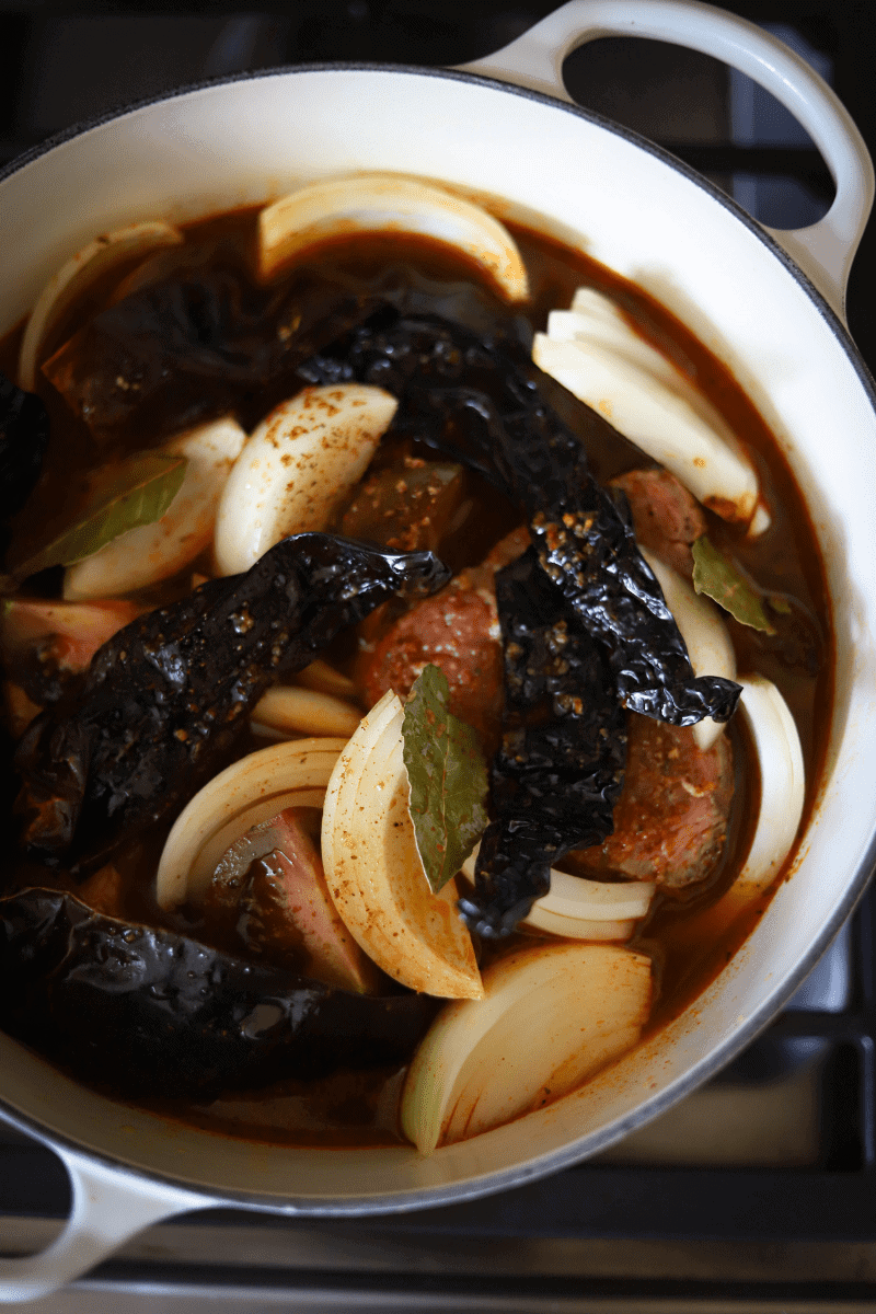 A soup pot filled with guajillo and pasilla peppers, beef roast, quartered onions and tomatoes in preparation for making Mexican birria