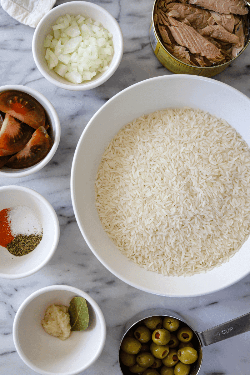 overhead photo of the main ingredients used to make Cape Verdean arroz de atum (tuna rice). Canned tuna, diced onions, quartered tomatoes, salt, pepper and seasonings, garlic, bay leaf and green olives