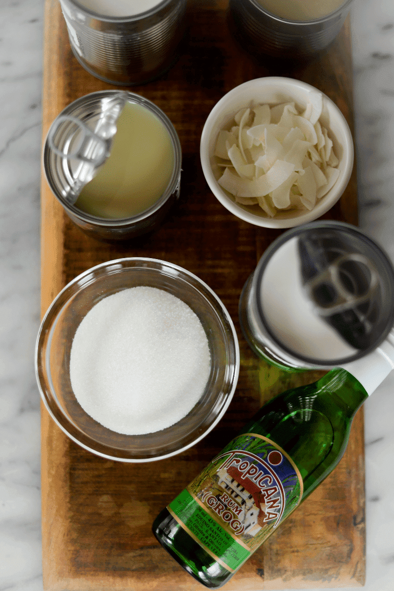 Overhead photo of all ingredients needed to make Cape Verdean ponche de coco (coconut ponche). Two cans of coconut milk, 1 can of condensed milk, granulated sugar, grogue and coconut flakes