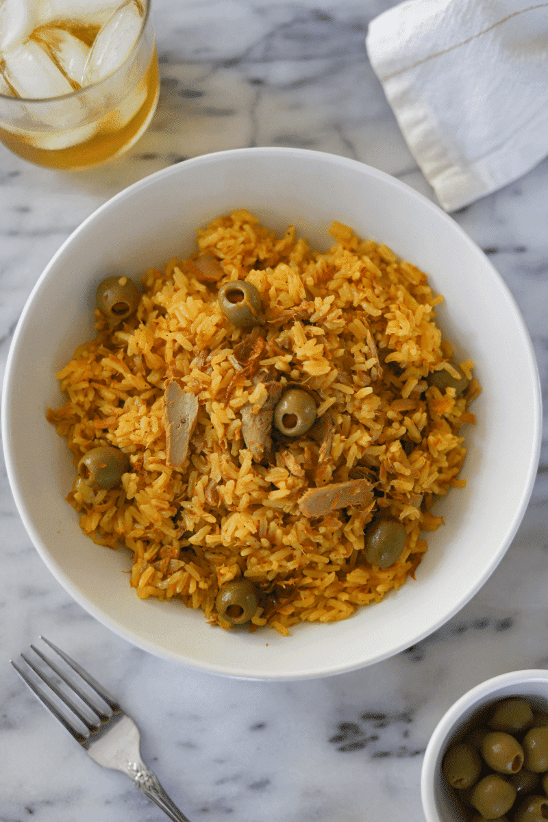 This is an overhead of Cape Verdean rice with tuna (arroz de atum) presented in a bowl with a napkin, beverage, fork and small bowl of olives off to the side.