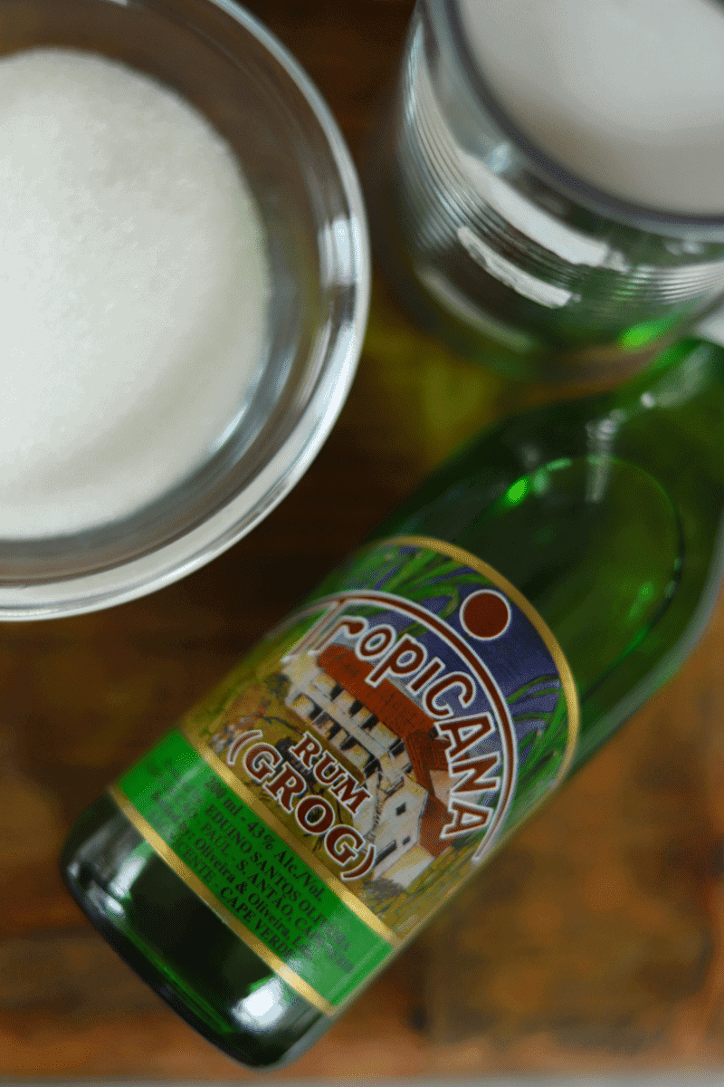 Close up photo of a bottle of Cape Verdean grogue laying down. There is a can of coconut milk and a small bowl of sugar in the frame.