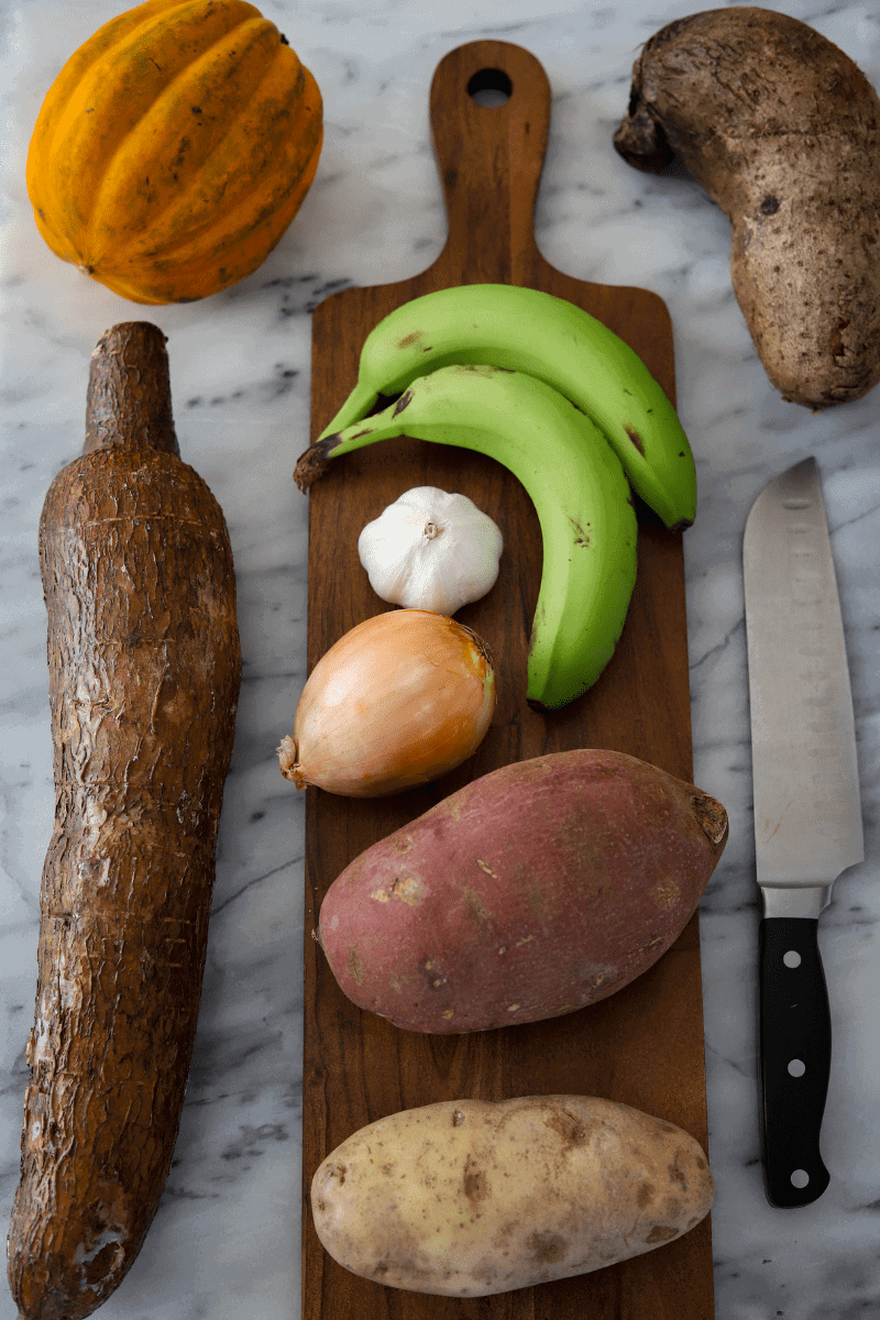 This photo demonstrates the produce and root vegetables needed to make modje (Cape Verdean goat stew). Yuca, acorn squash, green bananas, yam (inhame), onion, garlic, sweet potato (batata doce) and russet potato