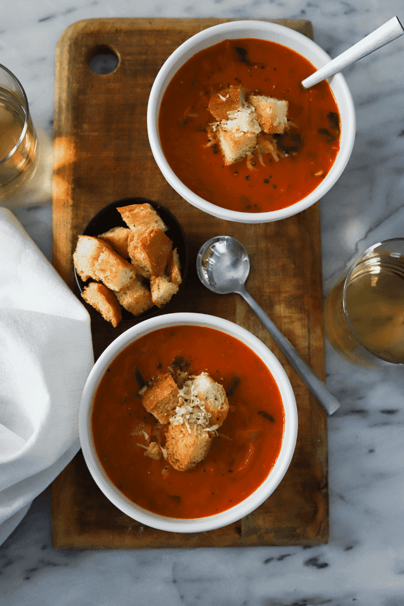 Two bowls of roasted tomato basil soup on a wood slab. Two glasses of wine, a small bowl of croutons and soup spoons are in the frame.