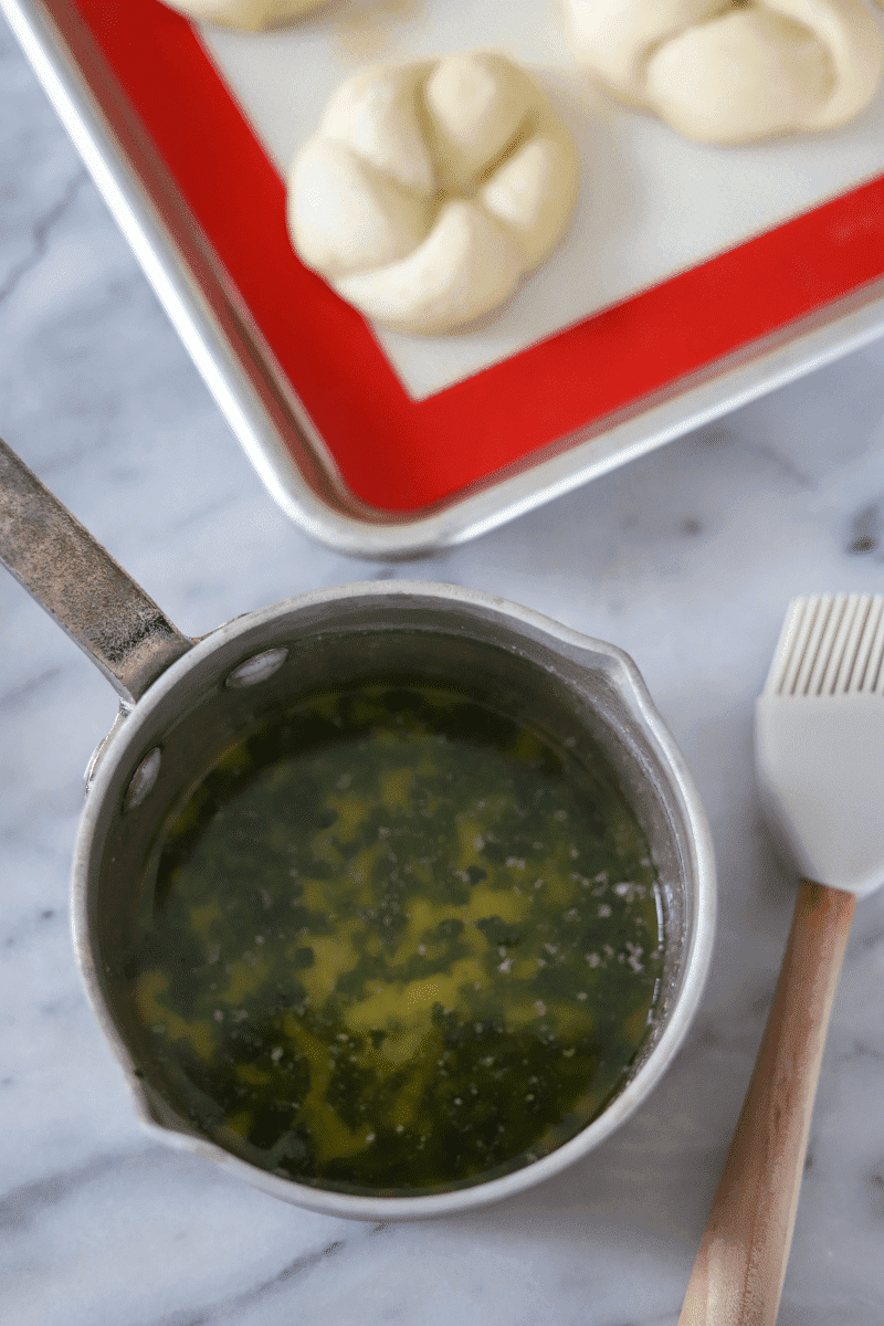 This is the melted butter with basil that is shown in a small saucepan, the unbaked garlic knots are arranged on a baking sheet in the background.