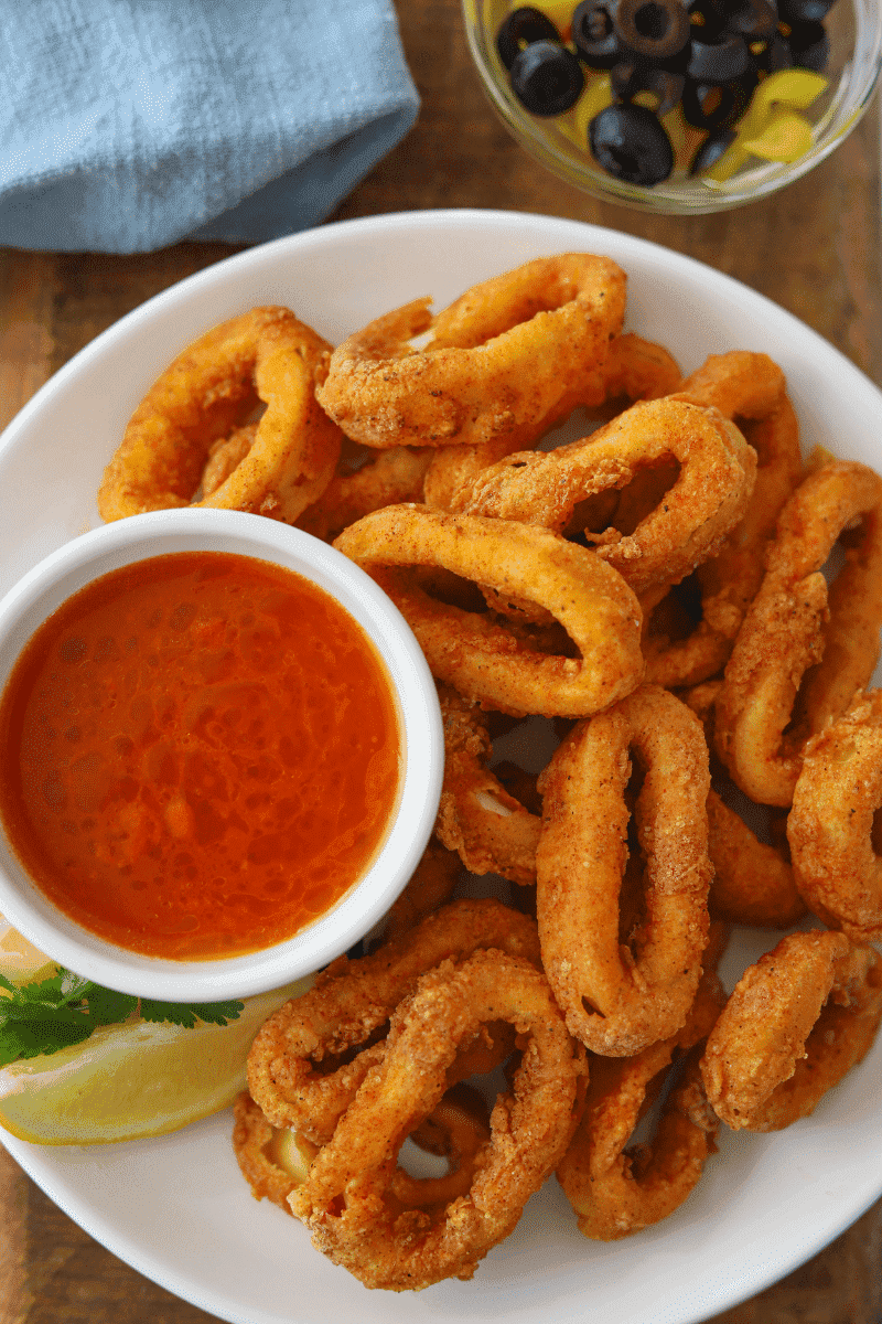 Portuguese style fried calamari (lulas fritas) on a white plate, garnished with fresh lemon, cilantro, banana peppers, olives and hot sauce