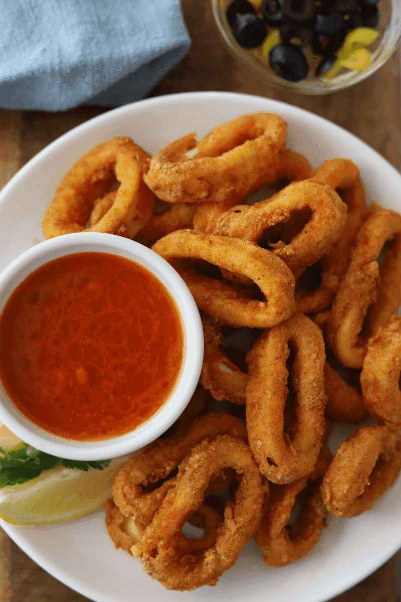 Portuguese style fried calamari (lulas fritas) on a white plate, garnished with fresh lemon, cilantro, banana peppers, olives and hot sauce