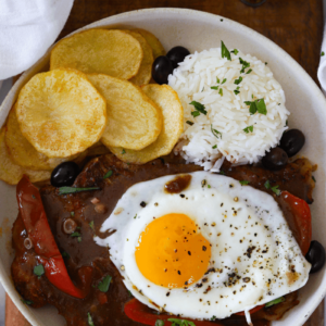 overhead shot of bitoque (portuguese steak) topped with egg and served with rice and fried potatoes