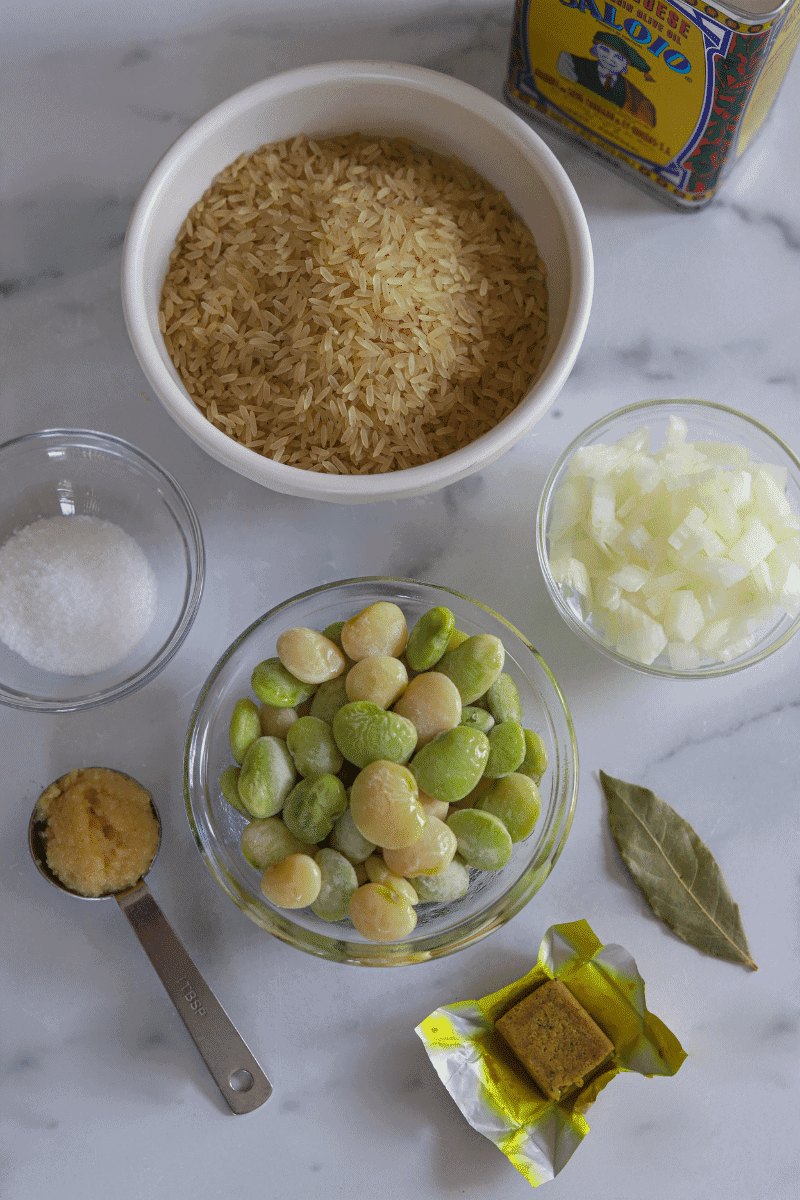 Overhead shot of ingredients for Cape Verdean style rice with lima beans: long grain white rice, extra-virgin olive oil, chopped onions, bay leaf, extra large bouillon cube, frozen fordhook lima beans, crushed garlic and salt