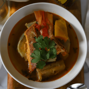 Close up of Cape Verdean caldo de peixe in a white bowl. Red snapper, squash, potatoes and banana are visible in the bowl. The dish is topped with a fresh sprig of cilantro.