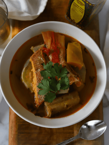 Close up of Cape Verdean caldo de peixe in a white bowl. Red snapper, squash, potatoes and banana are visible in the bowl. The dish is topped with a fresh sprig of cilantro.