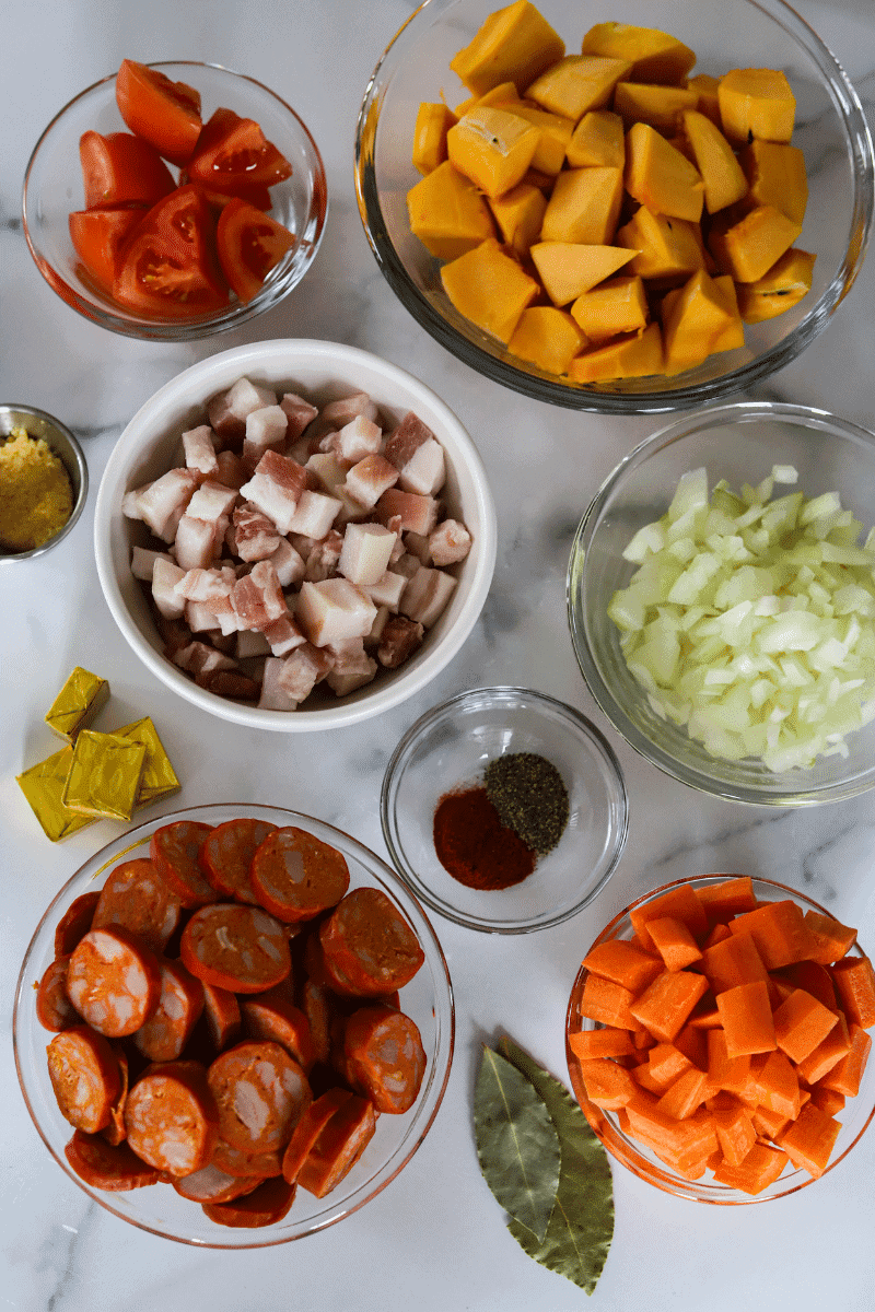 ingredients used for Cape Verdean feijoada - wedged tomatoes, cubed acorn squash, chopped onions, diced carrots, bay leaves, sliced chouriço, bouillon cubes, paprika, black pepper, diced salt pork, crushed garlic (beans are not shown)