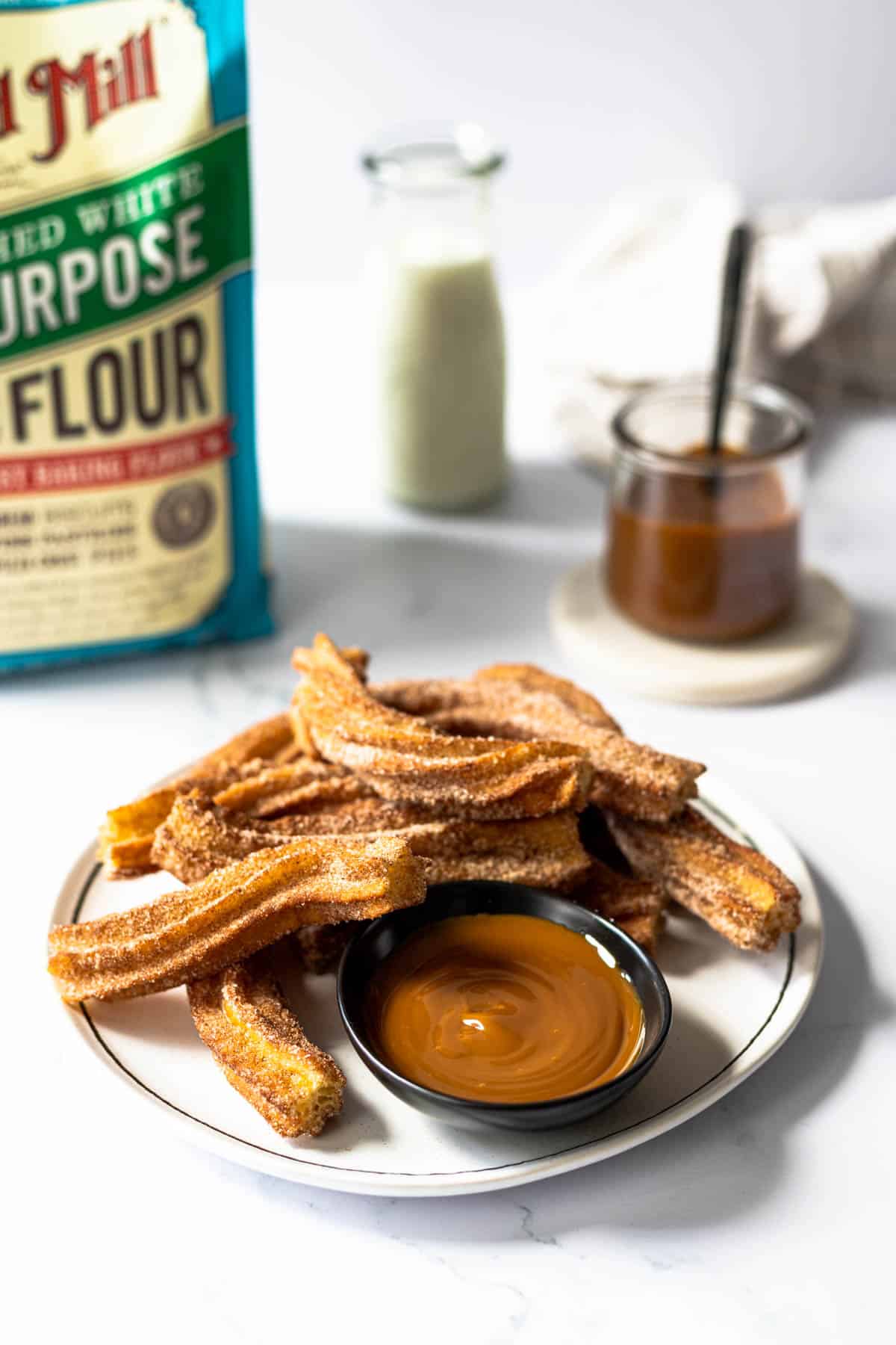 cinnamon sugar churros on a plate with dulce de leche for dipping. Bag of Bob's red mill flour in the background