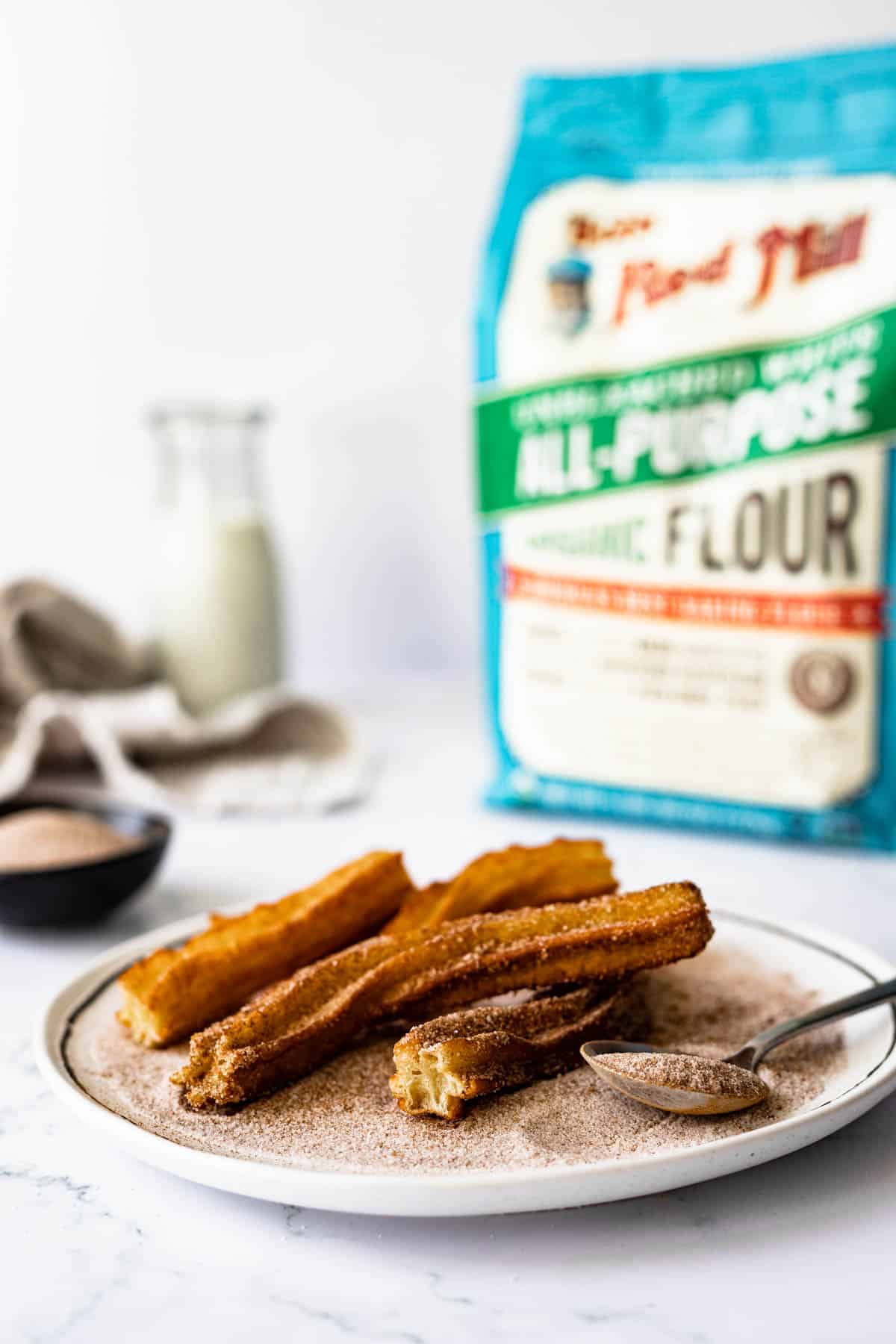 dusted churros on a plate of cinnamon/sugar - bag of Bob's Red Mill flour is shown in the background