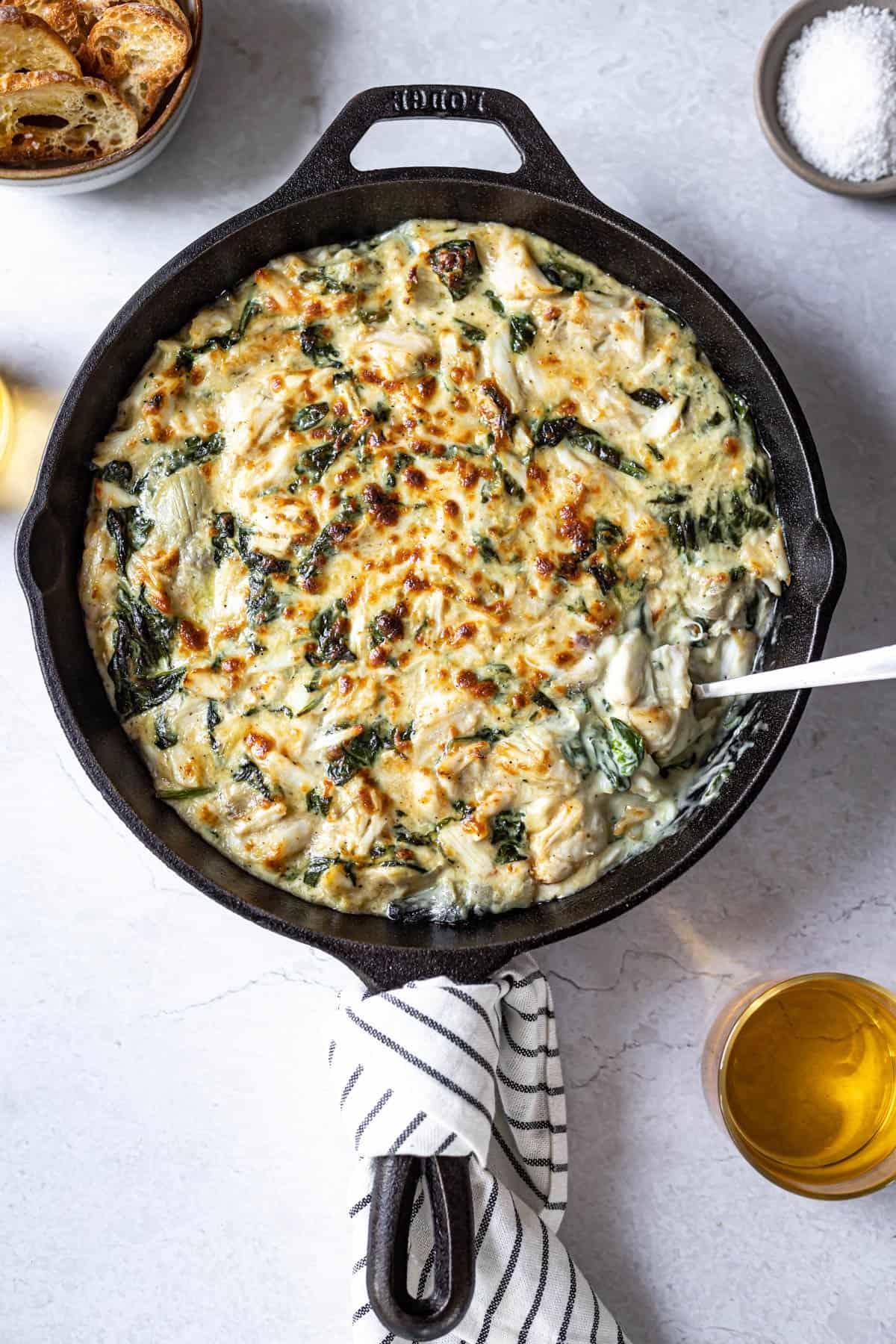 creamy, hot spinach crab and artichoke dip in a cast iron skillet served with crostini and glasses of wine