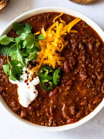 dutch-oven chili in a white bowl garnished with shredded cheddar cheese, fresh cilantro, sour cream, red chili flakes and sliced jalapeños.