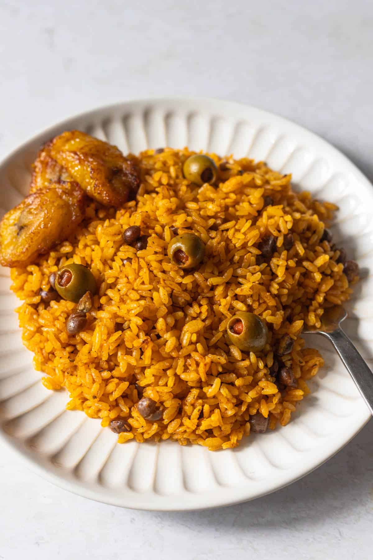 pigeon peas and rice (or arroz con gandules) on a white plate shown with green olives, a fork and side of sweet plantains.
