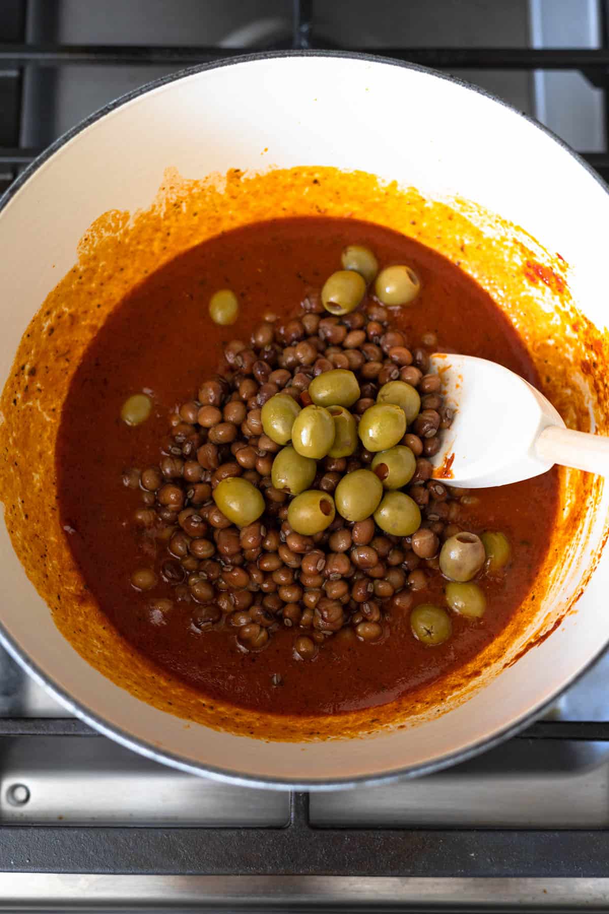 pigeon peas and olives in tomato-based sauce in a white dutch oven on the stove.