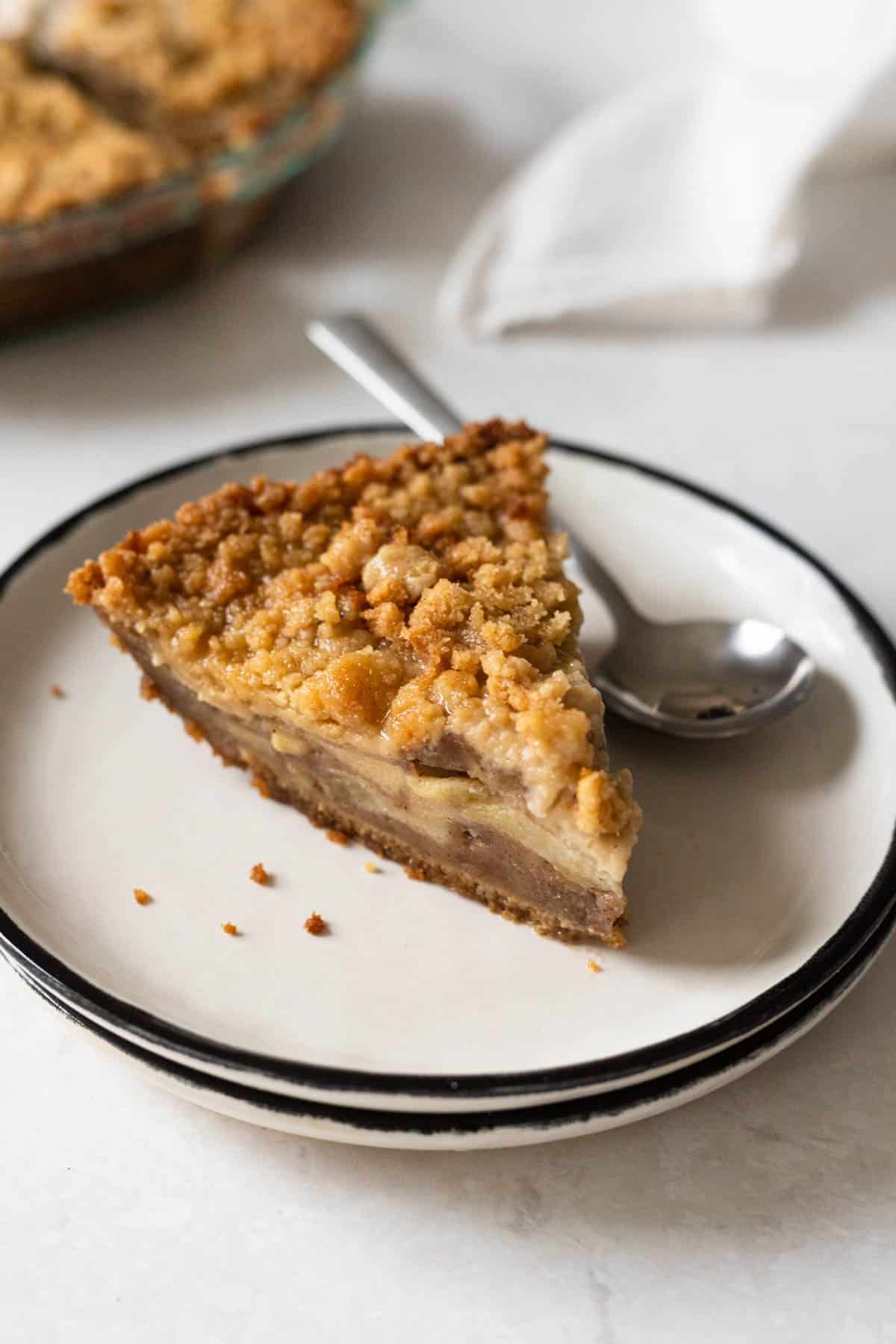 slice of apple pie with graham cracker crust on a stack of white plates. Whole pie in background.