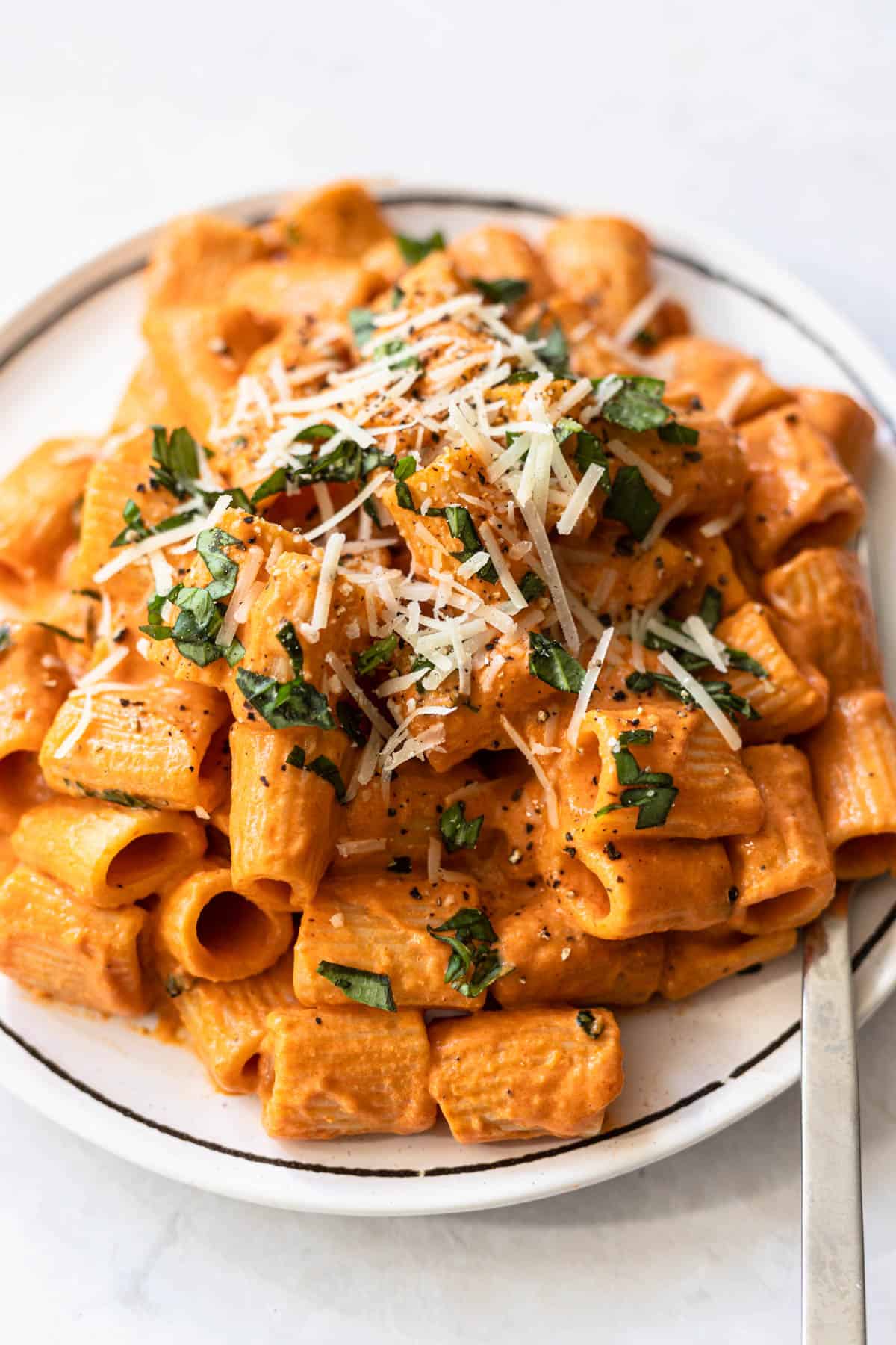 rigatoni tossed in pink sauce topped with shredded parmesan cheese and fresh basil on a white plate.